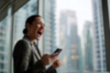 Fototapeta premium Blurred Businesswoman celebrating success with smartphone in modern cityscape