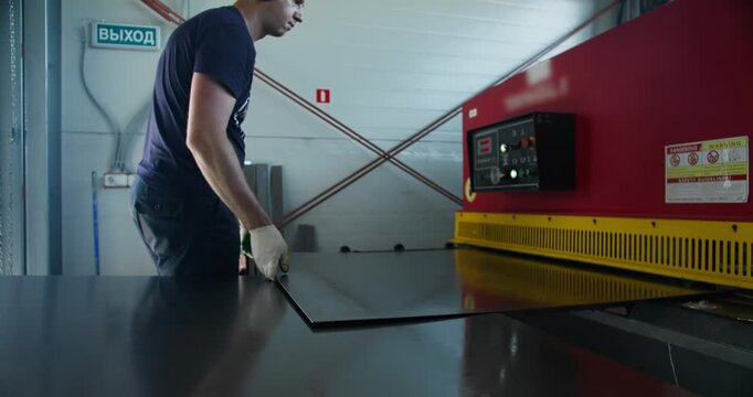 Worker operating metal cutting guillotine machine during industrial production process. Blue-collar labor and hands-on factory work in metal fabrication
