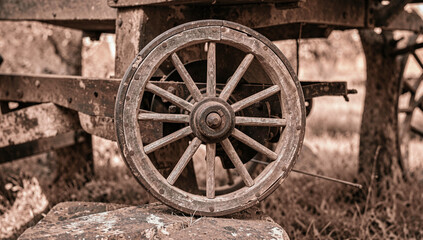 Fototapeta premium Weathered wooden wheel rests on stone. Rusty spokes show time's wear. Cart's sturdy frame leans slightly. Sepia tones evoke rustic charm. Background blurred, focusing on wheel