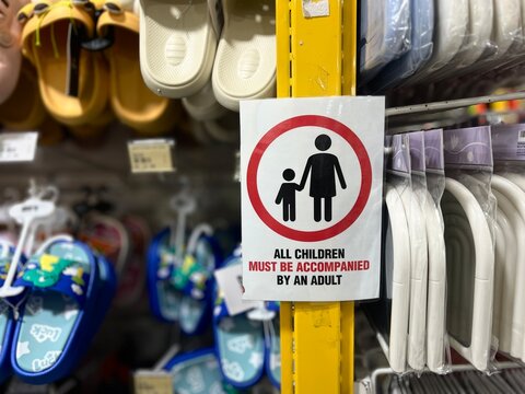 A retail safety warning sign stating all children must be accompanied by an adult posted on a yellow shelf in a department store.