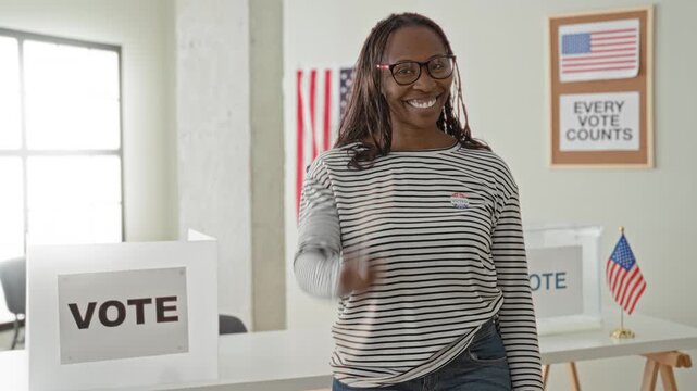 Woman points finger to camera in a polling building beside a vote box, sticker and american flag; civic pride.