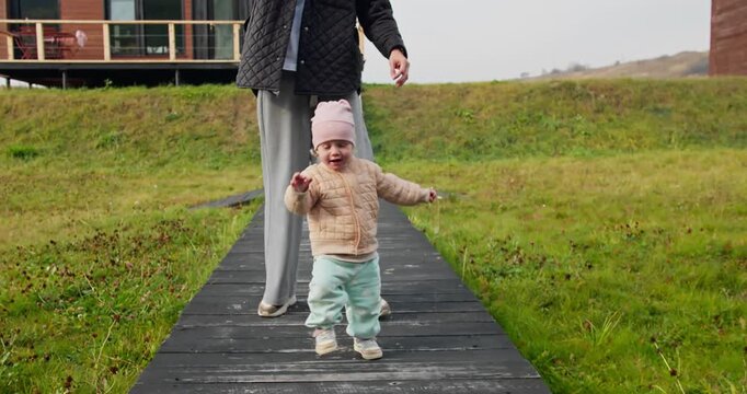 Toddler taking first steps on wooden path outdoors, early childhood moment with mother nearby in natural setting