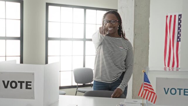 Woman giving thumbs up with hand beside ballot box and small us flag in a polling building wearing a voted sticker; civic pride.