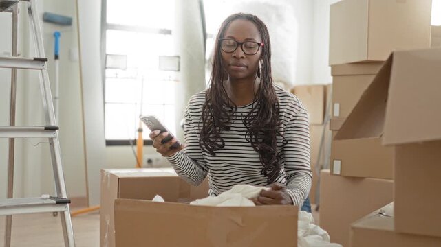 Woman holding smartphone while packing cardboard boxes and pulling a sheet from a box inside a building; calm transition focus.