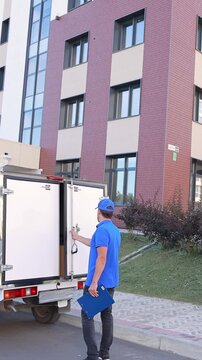 A male delivery man in a blue uniform opens a white cargo van and counts and checks boxes of goods