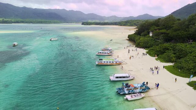 Drone shot flying along the white sandy shoreline of Kabira Bay, Ishigaki, featuring glass-bottom boats, tourists, and lush mountain ranges under an overcast sky.