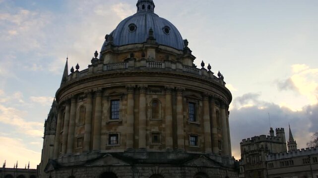 A low angle shot captures Oxford iconic Radcliffe Camera at dusk its grand dome and classical columns silhouetted against a soft blue sky with wispy clouds ideal for academic tourism or historic lifes