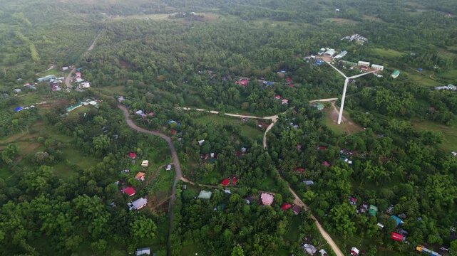 Aerial view of a wind turbine overlooking a tropical village with forested hills in Guimaras, Visayas, Philippines. Renewable energy infrastructure set within a rural landscape and lush greenery.
