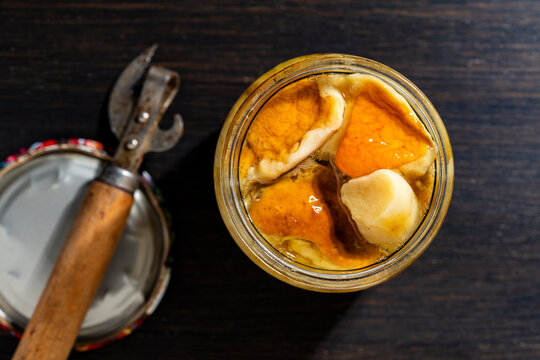 Marinated wild mushrooms in glass jar and can opener. Open jar of pickled porcini mushrooms in oil and vintage can opener on wooden background. Traditional homemade forest harvest preservation concept