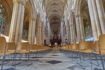 A long shot inside a grand cathedral, showing its vaulted ceilings, arched columns, stained glass windows, and rows of light wooden chairs in Beverley Minster - Yorkshire - Great Britain © Robert
