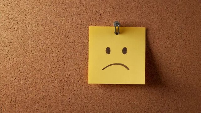 Sad face on yellow sticky note on corkboard background
