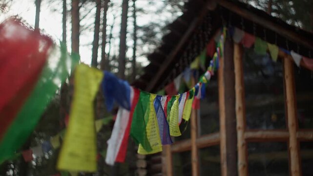 Close-up of vibrant Tibetan flags with blurred wooden house and pine trees in background. Cinematic bokeh and soft natural light