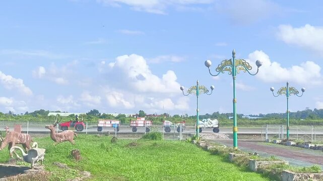 A small airport tug pulls luggage trailers along a runway behind a park decorated with animal statues and ornate lamp posts.