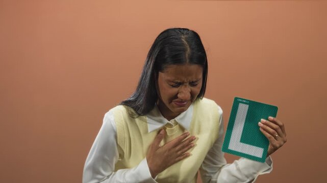 Woman clutching chest with one hand and coughing into a fist while holding a green learner l plate and looking pained in a studio set; anxiety.