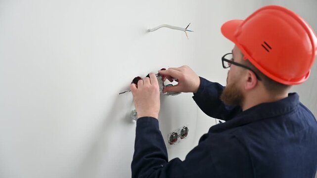 Electrician installs wall socket in unfinished interior. Worker in hard hat connects outlet mechanism during electrical wiring and home construction process