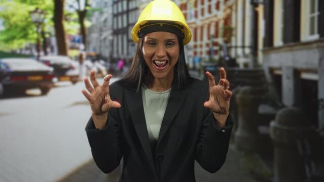 Woman architect in hardhat and blazer on street making clawing hands gesture with tongue out and broad smile before row of buildings; playful confidence.