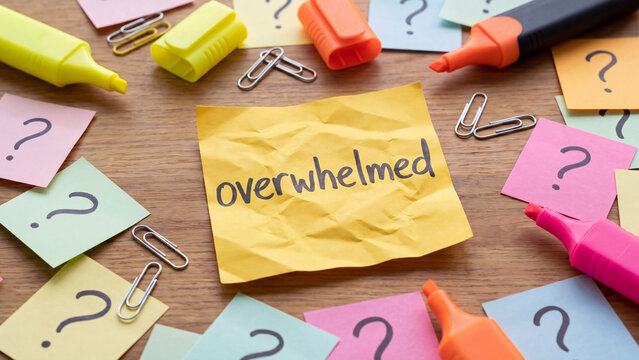 Overwhelmed workplace stress concept with crumpled sticky note surrounded by question marks highlighters and paperclips on cluttered desk for mental health anxiety and workload awareness