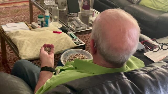 Anciano comiendo pipas y pelando c&aacute;scara con aud&iacute;fonos jubilado de tercera edad viendo la tele sentado en el sof&aacute; de espaldas en sal&oacute;n de casa