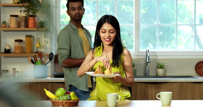 Indian husband walks into the kitchen and tries to grab a samosa from his wife who is holding a plate resulting in a friendly playful fight with laughter and joy in their modern home