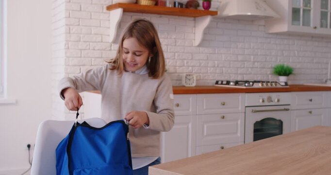 Joyful elementary school kid standing in kitchen, packing backpack with supplies before leaving. Happy child getting ready for day, feeling excited, motivated and prepared for learning routine.