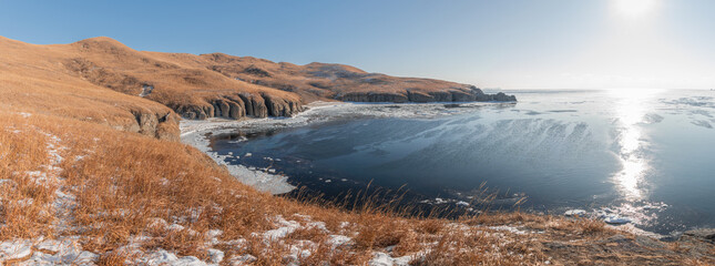 Naklejka premium Panoramic view of a winter landscape featuring a frozen lake surrounded by rocky cliffs and golden grass hills under a clear blue sky in a remote area