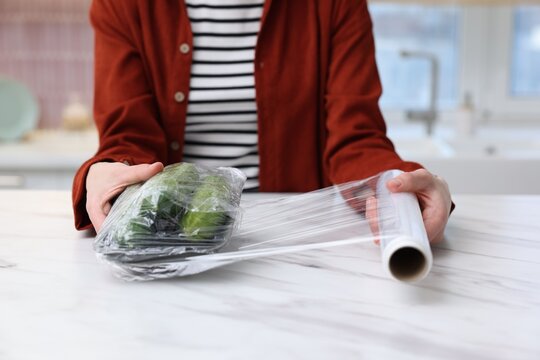 Woman wrapping container with fresh cucumbers in cling film at light marble table, closeup