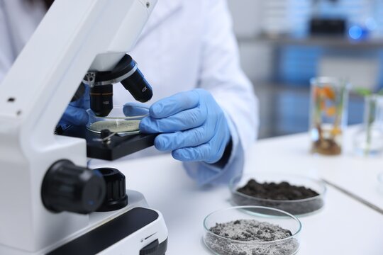 Laboratory testing. Scientist researching sand sample with microscope at white table indoors, closeup