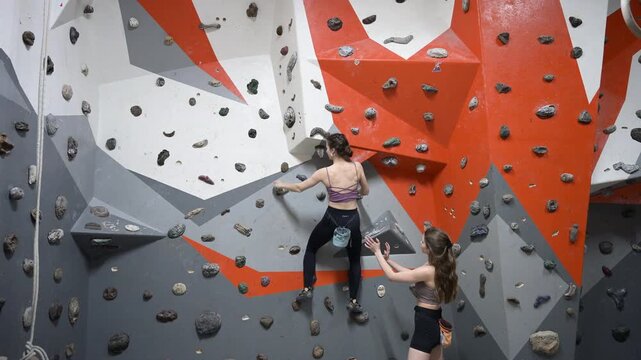 Female boulderer working on challenging route in gym while friend observes and spots. Stock video for sport performance, focus, resilience, and active recreation concepts