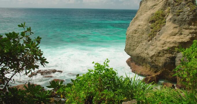 Turquoise ocean waves crashing against rugged limestone cliffs and green tropical plants