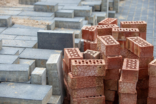 Stacks of heavy-duty gray concrete paving blocks and textured red perforated clay bricks arranged at a construction materials yard. Professional view of building and landscaping supplies ready for har