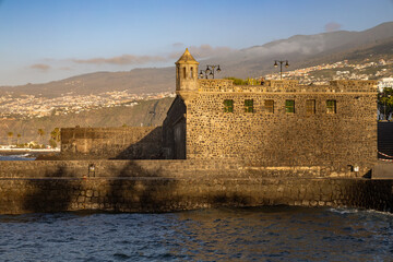 The historic stone watchtower and walls of the Batería de Santa Bárbara catch the warm evening light as waves crash against the base in Puerto de la Cruz, Tenerife © erikzunec