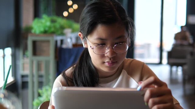 Young Asian student girl wearing glasses and earbuds using digital tablet for online learning and e-learning in a modern cafe atmosphere