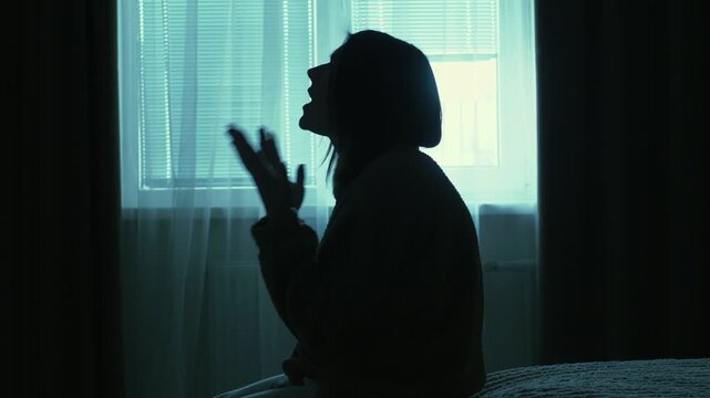 Young woman sitting alone in a dimly lit room, slowly raising hands in prayer near a window. Emotional scene of inner struggle, anxiety, loneliness and search for support, cinematic lighting and calm