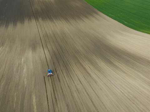 An aerial perspective of a blue tractor plowing a vast agricultural field in South Moravia, leaving behind a rhythmic pattern of dark furrows in the light brown earth