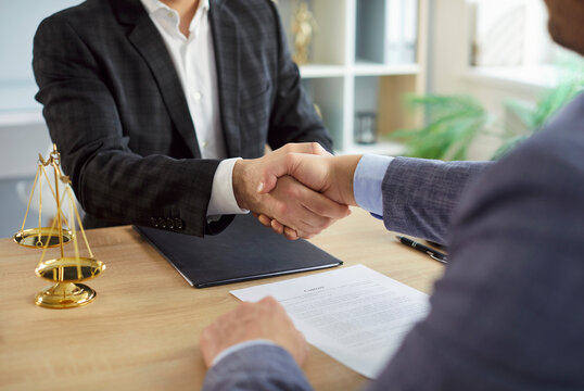 Close up photo of male lawyer in suit make a deal and shaking hand with a man client sitting at the desk on workplace in office with judge's scales on table. Legal law and advice concept.