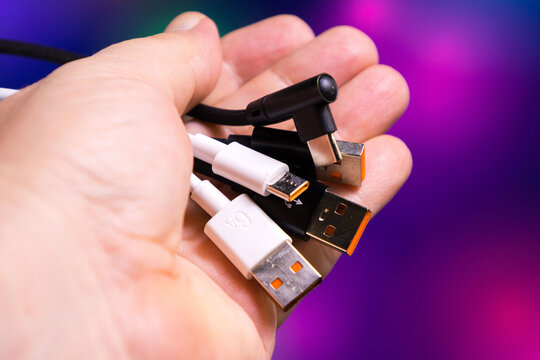 Multiple USB cables and chargers held in one hand showcasing various connector types including USB-A, USB-C, and an angled charger plug. Different cable designs and colors, such as black and white, ar
