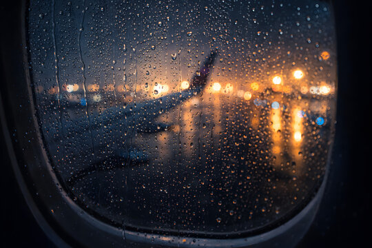 Close view of raindrops on airplane window with blurred runway lights outside, creating moody travel atmosphere and rainy weather concept.