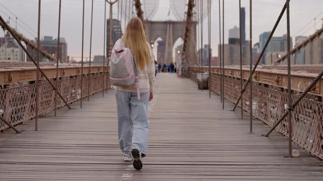 Young woman walking on Brooklyn Bridge with backpack and Manhattan skyline in New York