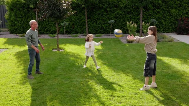 Active Grandfather Throws Ball While Playing With Two Grandchildren In Backyard