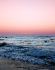 Obraz premium Pink sunset sky over calm blue ocean waves rolling onto sandy beach in Rethymno, Greece