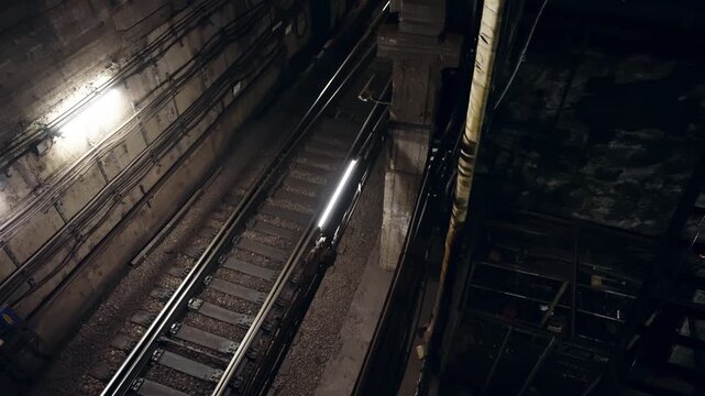 Dimly lit underground railway tunnel with parallel steel tracks, concrete pillars, and dense cabling, illuminated by fluorescent fixtures in an industrial urban transit corridor