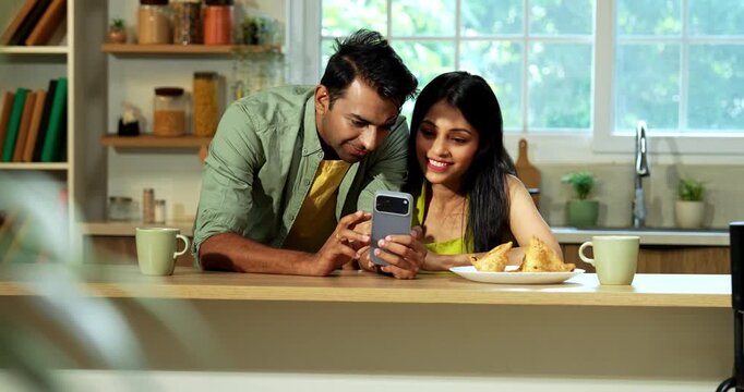 Indian asian young husband and wife browsing using smartphone in modern kitchen at platform surfing internet and checking photos wirelessly together during their weekend morning at home