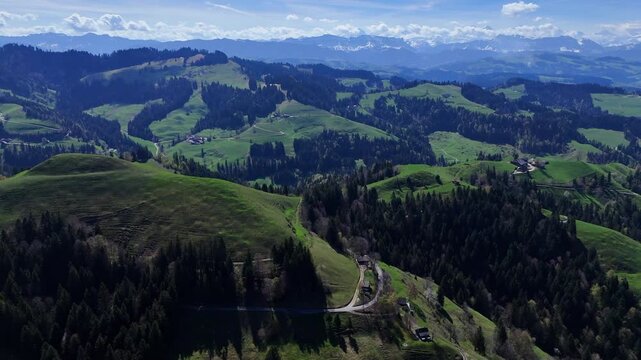 Wide aerial view of Swiss alpine foothills, patchwork meadows, dense woodland, and distant snowcapped mountains under blue sky.