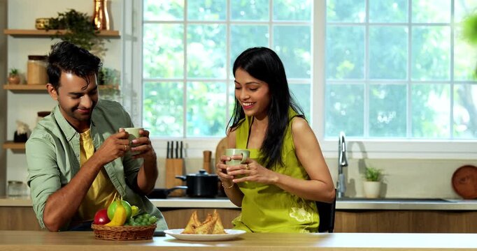 Indian asian young husband and wife having coffee in modern kitchen platform with light hearted chit chat enjoying their morning beverage and talking together in a relaxed home environment