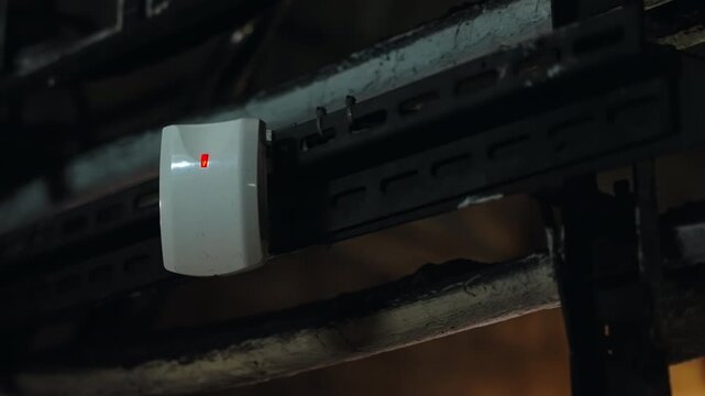 Closeup of a mounted security sensor with bright LED indicators on a metal support rail above weathered cable trays in a dim industrial corridor, signaling active monitoring
