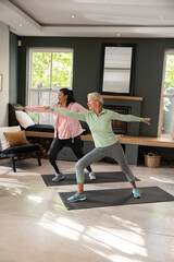 Naklejka premium Senior female friends practicing standing lunge on exercise mats in living room with mirror