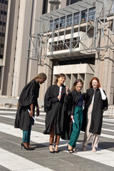 Diverse female graduates celebrating on campus plaza in gowns holding diplomas with red ribbons © wavebreak3