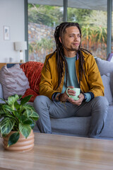 African American man wearing mustard coat sitting on sofa holding green mug looking out window