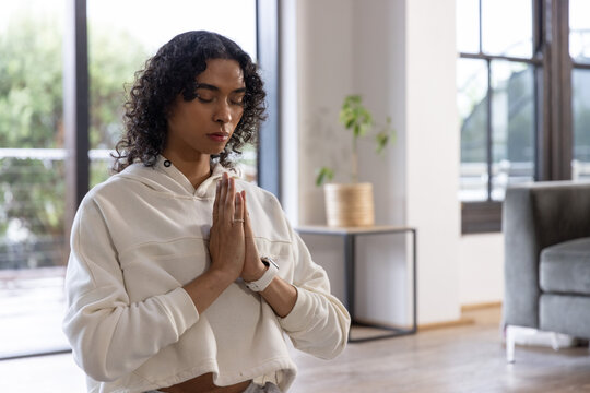 Non-binary adult meditating with hands in prayer at home on hardwood floor wearing cropped hoodie