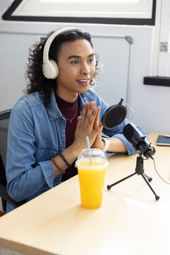 Non-binary creator speaking into microphone while wearing white headphones at desk with orange cup